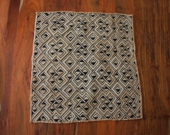 May include: Square beige and black patterned textile. The design features interlocking geometric shapes, creating a repeating pattern. The textile is placed on a wooden surface, showcasing its texture and design. The edges are finished with a simple border.