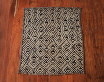 May include: Square textile with a geometric pattern in black and beige. The design features interlocking shapes, creating a repeating motif. The edges are finished with a dark brown border. The textile is placed on a wooden surface.