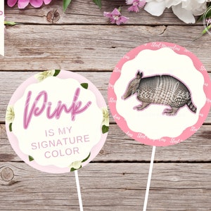 May include: Two pink and white round cake toppers. One topper features a pink floral design with the text "Pink is my signature colour". The other topper features a brown and white armadillo on a pink background.