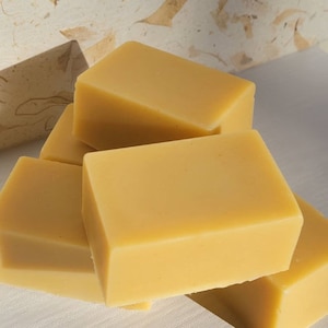 Puede incluir: Cuatro barras rectangulares de jabón amarillo apiladas una encima de la otra. El jabón es de color amarillo claro y tiene un acabado liso y mate.