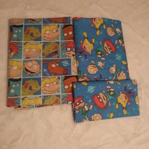 Puede incluir: Tres piezas de tela dobladas con personajes de dibujos animados. Una pieza tiene un patrón de cuadrícula con caras de personajes sobre un fondo azul claro. Las otras dos piezas tienen un fondo azul con personajes de dibujos animados y estrellas.