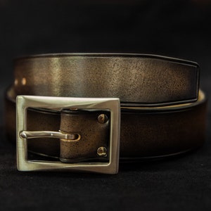 Peut inclure: Une ceinture en cuir marron avec une boucle argentée. La ceinture est pliée en deux et la boucle est au centre de l'image.