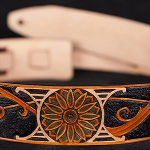 Puede incluir: Una correa de guitarra de cuero negro con un dise&ntilde;o floral detallado en naranja, verde y blanco. La correa tiene una hebilla y un extremo de correa.