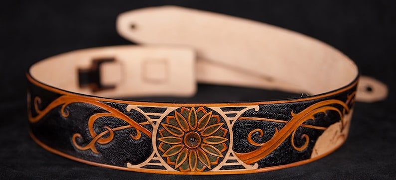 Puede incluir: Una correa de guitarra de cuero negro con un dise&ntilde;o floral detallado en naranja, verde y blanco. La correa tiene una hebilla y un extremo de correa.