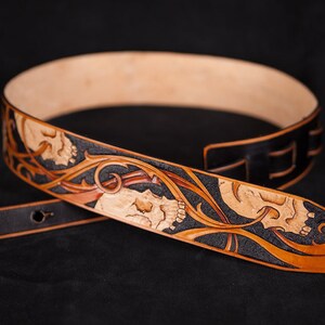Puede incluir: Una correa de guitarra de cuero marr&oacute;n con fondo negro y una talla detallada de calaveras y vides.
