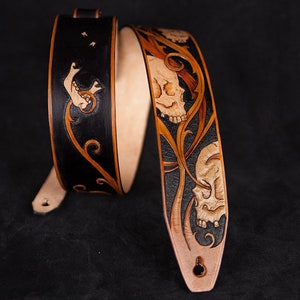 Puede incluir: Una correa de guitarra de cuero negro y marr&oacute;n con un dise&ntilde;o de calavera y flores. La correa est&aacute; decorada con intrincados tallados y es un accesorio &uacute;nico para cualquier m&uacute;sico.
