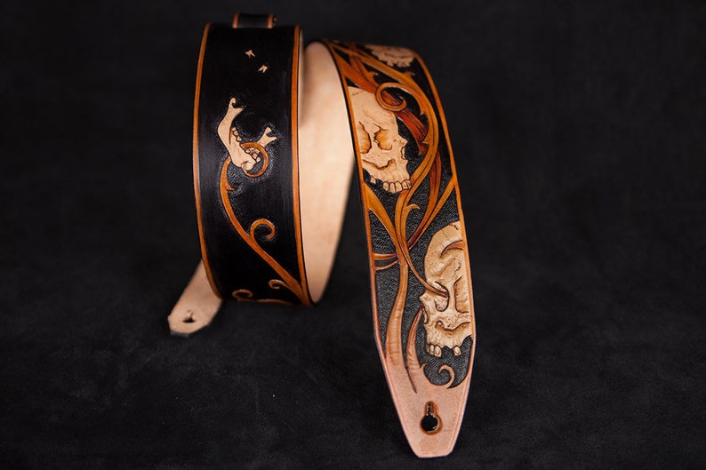 Puede incluir: Una correa de guitarra de cuero negro y marr&oacute;n con un dise&ntilde;o de calavera y flores. La correa est&aacute; decorada con intrincados tallados y es un accesorio &uacute;nico para cualquier m&uacute;sico.