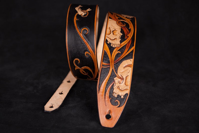 Puede incluir: Una correa de guitarra de cuero negro y marr&oacute;n con un dise&ntilde;o de calavera y flores.