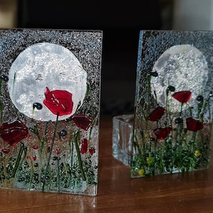 May include: Two glass bookends with a fused glass design. Each bookend features a full moon, red poppies, and green grass. The background is a textured, speckled grey. The bookends are rectangular and stand upright.