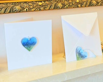 May include: Two white greeting cards, one with a blue, green, and glitter heart pendant attached. The other card is an envelope with a matching heart pendant. The pendants are attached with a silver-colored loop.