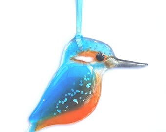 May include: A decorative glass ornament in the shape of a kingfisher bird. The bird features a blue body, orange belly, and a long, dark beak. A light blue ribbon is attached for hanging.
