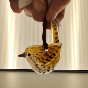 May include: A glass bird ornament with a yellow and brown speckled design. The bird has a dark brown beak and a loop for hanging. The ornament is suspended by a dark brown ribbon.