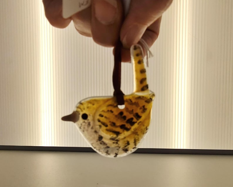 May include: A glass bird ornament with a yellow and brown speckled design. The bird has a dark brown beak and a loop for hanging. The ornament is suspended by a dark brown ribbon.