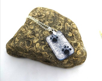 May include: A rectangular pendant necklace with a silver chain. The pendant has a blue and silver textured background with two black paw prints. The necklace is displayed on a textured, brown rock.