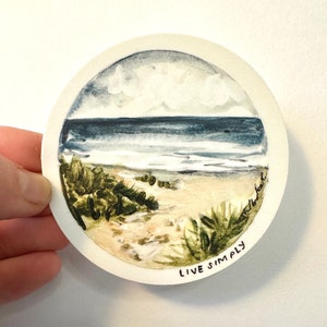 以下が含まれることがあります： ビーチの風景を描いた水彩画の丸いステッカーです。絵には青い海、白い波、砂浜、緑の茂みが描かれています。ステッカーの下部には「LIVE SIMPLY」という文字が印刷されています。
