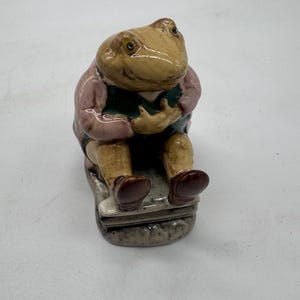 May include: A ceramic figurine of a seated toad wearing a pink jacket, a green vest, and brown shoes. The toad is positioned on a small, textured gray base, with its hands resting on its chest. The figure features a glossy, glazed finish.