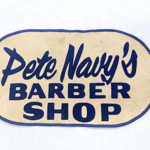 May include: A vintage oval-shaped sign with blue stitching around the edges. The sign is made of beige fabric and features the text "Pete Navy's Barber Shop" in blue lettering.