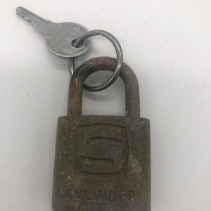 May include: A rusty padlock with a key attached. The padlock has a square shape with the word "CYLINDER" engraved on the bottom and a stylized letter "S" in the center. The key is silver and has the word "STARTER" engraved on it.