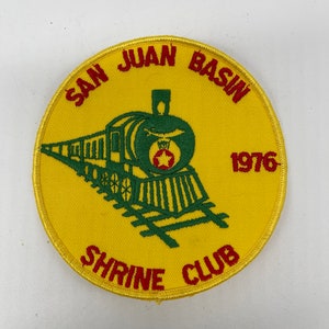 May include: A yellow circular patch with red embroidered text that reads "San Juan Basin Shrine Club 1976". A green embroidered train is in the center of the patch.