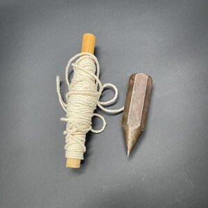 May include: A wooden spool of white string and a metal plumb bob, both tools used for measuring levels and angles in construction.