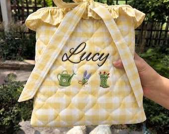 Può includere: Una borsa trapuntata a quadretti vichy gialli e bianchi con coulisse, che riporta il nome Lucy ricamato in corsivo nero. Sotto il nome sono presenti ricami raffiguranti un annaffiatoio verde, un rametto di lavanda viola e uno stivale da giardino.