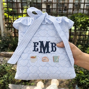 Pode incluir: Uma bolsa de seersucker azul claro e branco com textura acolchoada de corações e borda superior com babados. A bolsa apresenta um monograma bordado em azul escuro com as letras EMB sobre pequenos ícones bordados de uma caneca, um gato e um caderno.