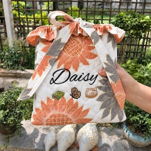 Pu&ograve; includere: Una pochette in tessuto trapuntato con sfondo bianco e motivi di girasoli arancioni e grigi. La pochette presenta un bordo superiore arricciato, una chiusura a laccio ed &egrave; ricamata con il nome Daisy, una piccola farfalla e due icone.