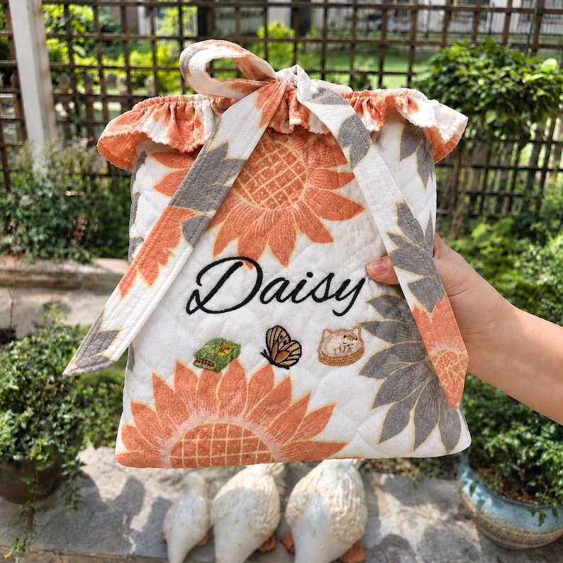 Pu&ograve; includere: Una pochette in tessuto trapuntato con sfondo bianco e motivi di girasoli arancioni e grigi. La pochette presenta un bordo superiore arricciato, una chiusura a laccio ed &egrave; ricamata con il nome Daisy, una piccola farfalla e due icone.
