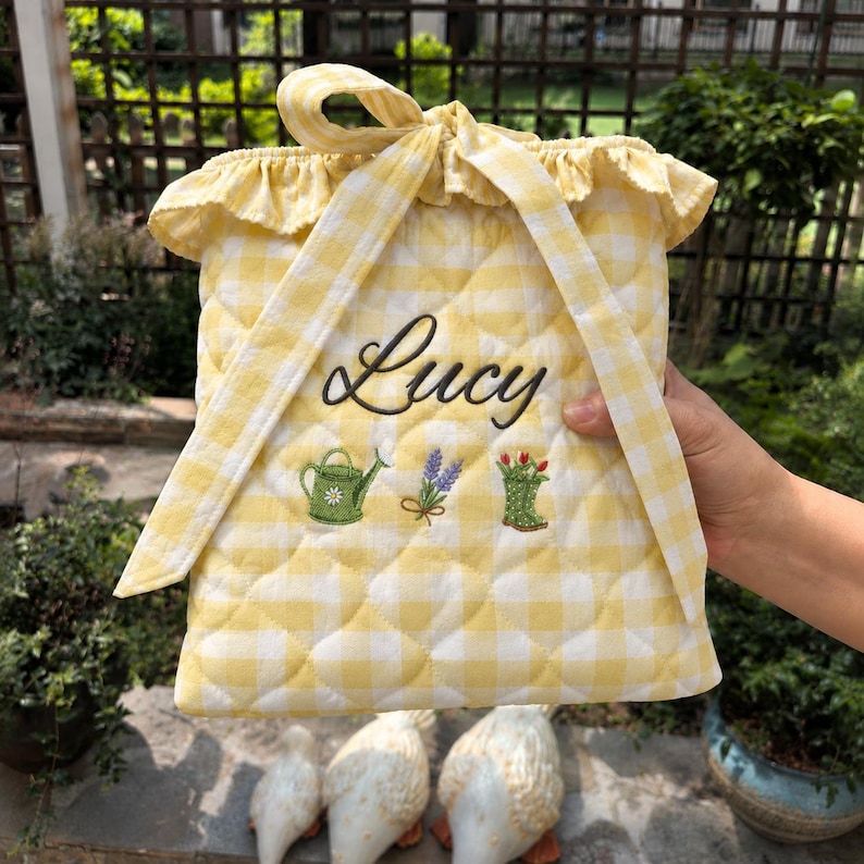 Pu&ograve; includere: Una borsa trapuntata a quadretti vichy gialli e bianchi con coulisse, che riporta il nome Lucy ricamato in corsivo nero. Sotto il nome sono presenti ricami raffiguranti un annaffiatoio verde, un rametto di lavanda viola e uno stivale da giardino.