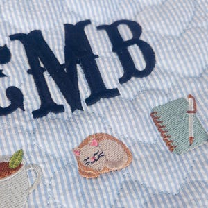 Pu&ograve; includere: Un tessuto trapuntato a righe sottili azzurre e bianche con le lettere EMB ricamate in blu scuro. Il tessuto presenta piccoli motivi ricamati raffiguranti una tazza grigia con una foglia verde, un gatto beige che dorme e un taccuino verde chiaro.