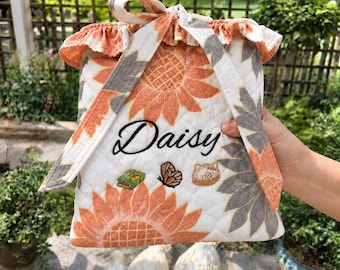 Può includere: Una pochette in tessuto trapuntato con sfondo bianco e motivi di girasoli arancioni e grigi. La pochette presenta un bordo superiore arricciato, una chiusura a laccio ed è ricamata con il nome Daisy, una piccola farfalla e due icone.