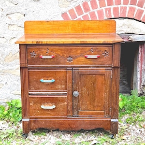 May include: A wooden cabinet with a rectangular top, featuring one wide top drawer with carved floral details, two smaller drawers below it, and a single side door. The drawers have colorful metal handles, and the door has a round knob.