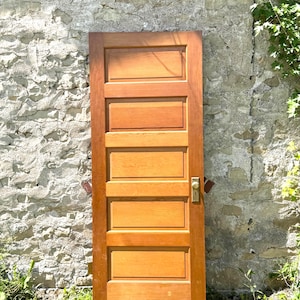 May include: A rectangular wooden door featuring five horizontal recessed panels of equal size. The door has a warm, medium-brown wood grain finish and includes a brass-colored metal door knob and a rectangular escutcheon plate on the right side.