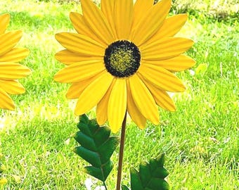 May include: A metal sunflower garden stake featuring bright yellow petals with a textured black and yellow centre. The flower is attached to a thin brown stem with two jagged green leaves extending from the sides, creating a floral garden ornament.