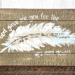 May include: A rectangular wooden sign made of three horizontal planks with a weathered brown finish. A large white feather is painted in the center, with the white text "God gave me you for the ups and downs" written across the top and bottom.