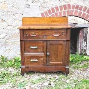 May include: A vintage wooden cabinet with a light brown top and dark brown body. The cabinet has two drawers with colorful handles and a single door. The cabinet is on wheels and has decorative floral carvings.