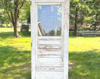 Puede incluir: Una puerta de madera alta y rectangular con un panel de vidrio en la parte superior y tres paneles horizontales empotrados debajo. La puerta tiene un acabado de pintura blanca desgastada, veta de madera visible y bisagras de metal en el lateral.