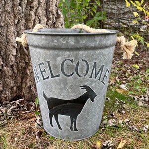 May include: A galvanized metal bucket with a rope handle and the word "WELCOME" printed on the side. A black silhouette of a goat is also printed on the bucket.