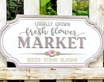 May include: A rectangular metal sign with rounded edges featuring the text LOCALLY GROWN fresh flower MARKET in grey and brown lettering. A small pink flower graphic sits above the words SEEDS STEMS BLOOMS in a light brown font.