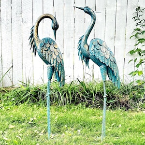 May include: Two metal heron garden statues featuring a teal and gold patina finish. Each bird stands on a single twisted metal stake. One heron has its head bowed downward, while the other heron stands with its head held upright and alert.