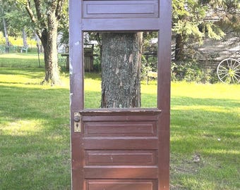 Puede incluir: Una puerta de madera de color marrón oscuro con una gran abertura rectangular de cristal en la parte superior, tres paneles horizontales empotrados en la parte inferior y una placa de pomo metálica en el lado izquierdo del marco.