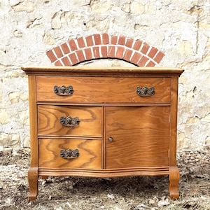 May include: A light brown antique wooden cabinet with two drawers, two smaller drawers, and a cabinet door. The cabinet has ornate bronze-colored handles and sits on curved legs. A brick arch is above the cabinet.