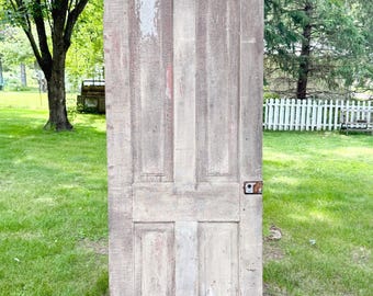 Puede incluir: Una puerta de madera antigua con cuatro paneles, que muestra signos de desgaste y pintura desgastada. La puerta está apoyada contra un árbol en un entorno exterior cubierto de hierba. Una alfombra de bienvenida está colocada en la base de la puerta.