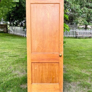 May include: A wooden door with a warm, light brown finish, featuring a rectangular panel design. The door has a brass doorknob and is set against a backdrop of green grass and trees. A black welcome mat is at the base.