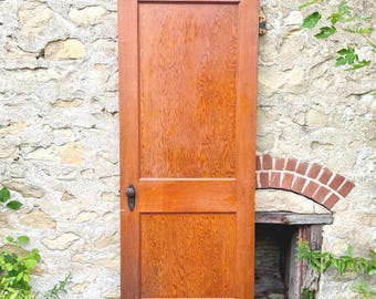 May include: A rectangular wooden door with a warm brown finish and a visible wood grain pattern. The door features two vertical panels, one larger top panel and one smaller bottom panel, and includes a dark metal round knob on the left side.