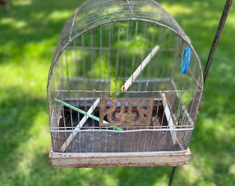 May include: A rusty metal birdcage with a white base hangs from a black metal stand. The cage has a decorative metal design on the front and two wooden dowels inside.
