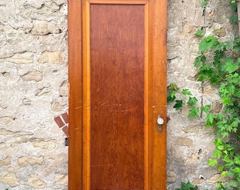 May include: A rectangular wooden door with a medium brown finish, featuring a single large central panel with a visible wood grain pattern. The door includes a round glass doorknob and a metal keyhole plate mounted on the right side frame.