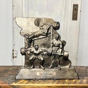 May include: A bronze-colored trophy with three figures of weightlifters. The trophy has a rectangular base with a plaque that reads "Mid-Mosa Tn Open 360 Level 2 Allen Powerlifting".