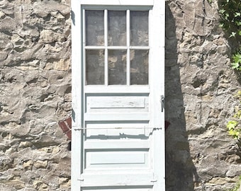 Puede incluir: Una puerta de madera pintada de gris claro con una ventana de seis paneles en la parte superior y cuatro paneles horizontales empotrados debajo. La puerta incluye un mecanismo de pestillo de metal en el lado derecho y una varilla metálica fina.