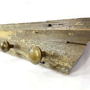 May include: A rustic wooden coat rack with three brass knobs. The wood has a weathered appearance with peeling paint, showcasing a blend of gray, brown, and yellow tones. The knobs are evenly spaced and add a touch of vintage charm.
